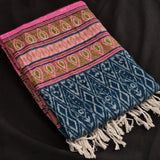 A folded woven textile throw blanket with cream fringe, featuring a pink and olive green patterned upper section and a blue lower section with white geometric motifs, laid on a black background.