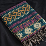 A folded woven throw blanket with a Southwestern geometric pattern in teal, mustard yellow, pink, and cream, finished with fringed edges, displayed on a black fabric background.