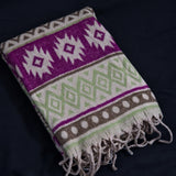 A folded woven throw blanket with a Southwestern geometric pattern in purple, sage green, brown, and cream, featuring fringed edges, displayed on a dark navy background.