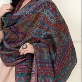 A woman modeling a large, multicolored patterned shawl draped over her shoulders, paired with a light pink dress against a white paneled wall background.