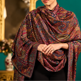 A model is shown wearing a long, open-front shawl or lightweight coat with an intricate multicolor paisley and floral pattern in deep red, blue, green, and gold tones, set against a dark teal background with ornate gold furniture.