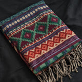 A folded woven throw blanket with a colorful geometric pattern and fringed tassels, laid on a dark fabric background.