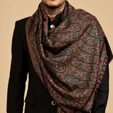 A split-screen studio photo showing a male model wearing a large paisley shawl scarf in black over a black outfit, against a solid beige background.