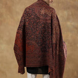 A man wearing glasses is shown from the back, draped in a large dark red and black patterned shawl over a black kurta and white trousers, standing against a textured brown studio backdrop.