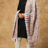A man modeling a light pink and white patterned shawl with multicolor borders over a black kurta, white trousers, and black loafers, set against a textured brown studio backdrop.