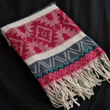 A folded woven throw blanket with fringe, featuring a Southwestern geometric pattern in red, cream, slate blue, and teal on a black fabric background.