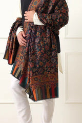 A man modeling a long, multicolored patterned shawl draped over a black kurta and white trousers, standing against a cream-colored paneled wall.