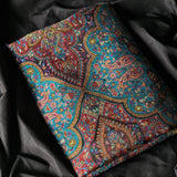 A folded patterned textile with an intricate paisley and floral design in turquoise, red, purple, pink, and gold, laid on a dark black fabric background.
