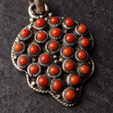 A close-up of a silver-toned pendant featuring a cluster of round, bright orange-red cabochon stones set in a textured, rope-bonded silver border, shown on a dark textured background.