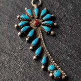 A silver-toned Native American style cross pendant decorated with turquoise blue teardrop stones and a central red stone, shown on a dark textured slate background.