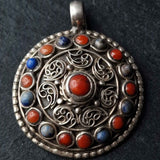 An ornate circular silver pendant featuring intricate filigree patterns and a central red coral cabochon, surrounded by a border of alternating red and blue coral stones, set against a dark textured background.