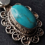 A close-up of a silver-toned pendant featuring a large oval turquoise stone with natural matrix lines, set in an ornate filigree-style frame against a dark background.