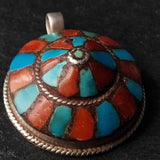 A round silver pendant featuring a colorful inlay pattern of turquoise, coral, and red stones, photographed on a dark textured background.