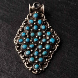 An ornate silver-toned pendant featuring a cluster of small turquoise blue stones set in a detailed filigree frame, photographed on a dark textured background.
