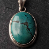 An oval turquoise pendant with a silver bezel setting, displayed on a dark textured slate background.
